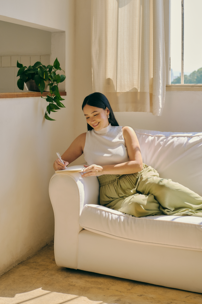 An image of a woman sitting down on a couch and writing in a book.