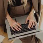 Image of a woman having a laptop on her lap whilst sitting on a desk and wearing a brown suit with a black blouse underneath.