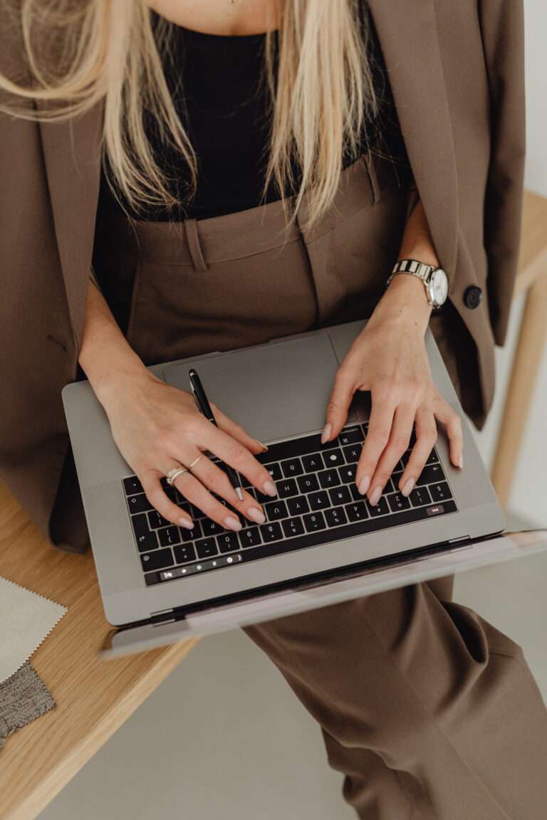 Image of a woman having a laptop on her lap whilst sitting on a desk and wearing a brown suit with a black blouse underneath.