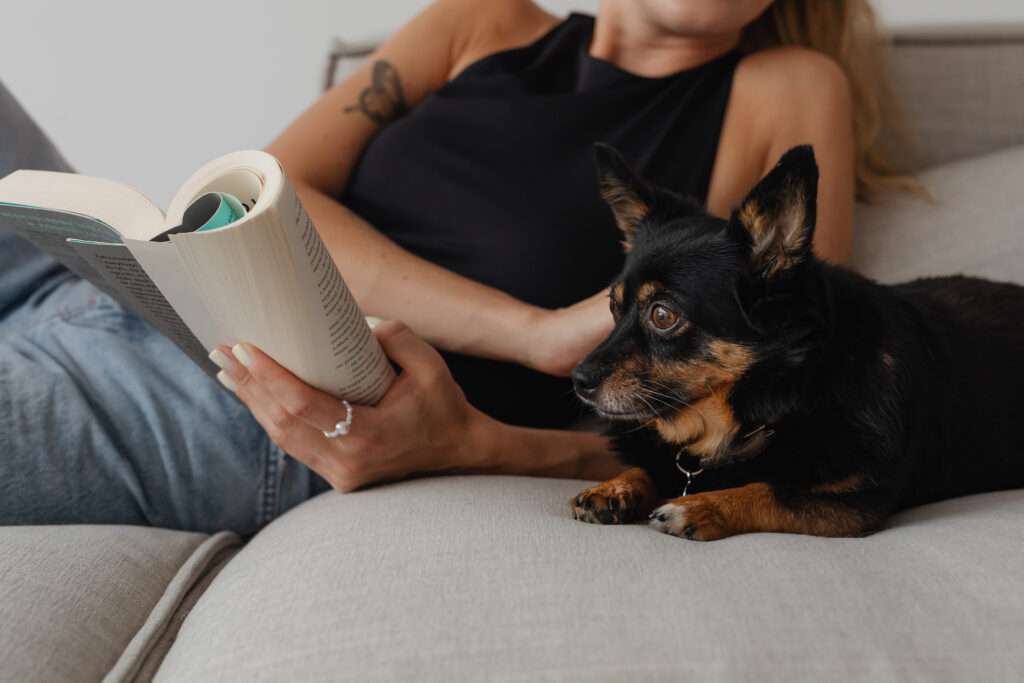 A woman reading a book on the sofa with a dog next to her.
