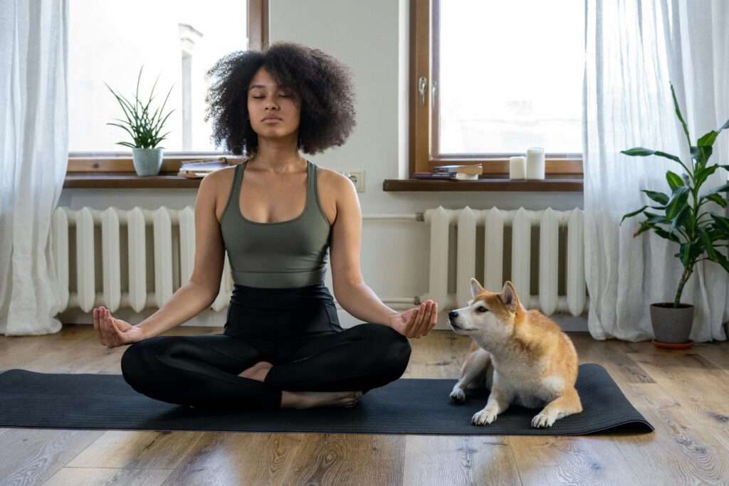 Woman on a yoga mat in her living room meditating with her dog beside her