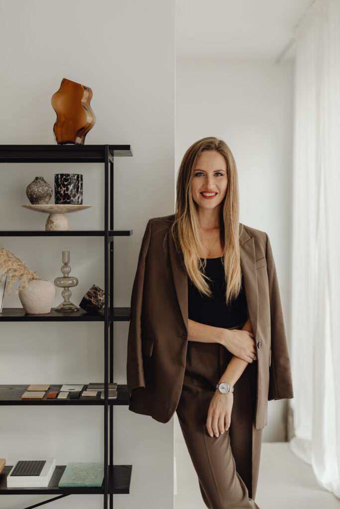 A woman wearing a brown suit, standing next to a shelf, and smiling.
