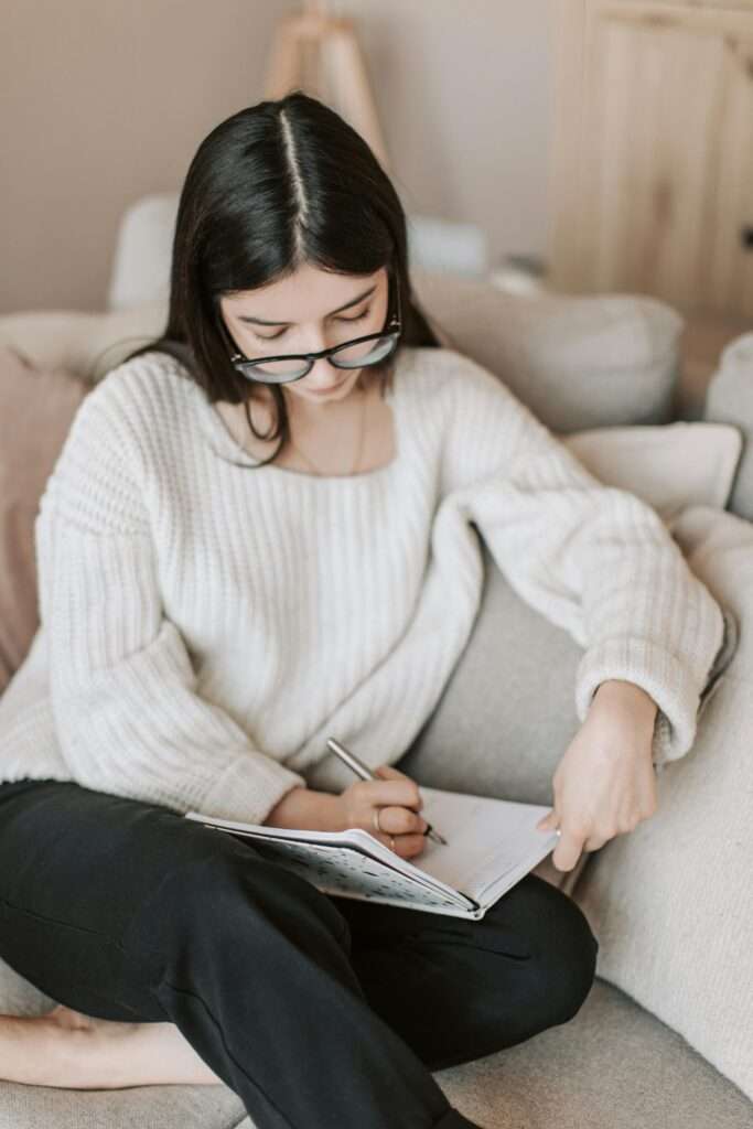 A woman writing in a notebook while sitting on a couch.
