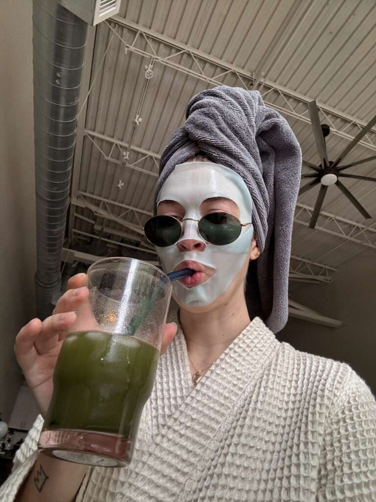 Image of a woman wearing a robe, sunglasses, towel head wrap, facial mask, and drinking a glass of green juice.