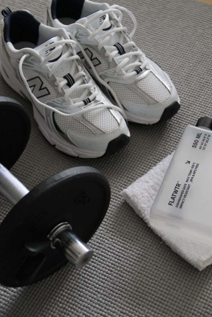Image of a New Balance tennis shoes, a dumbbell, and white towel on a carpet.