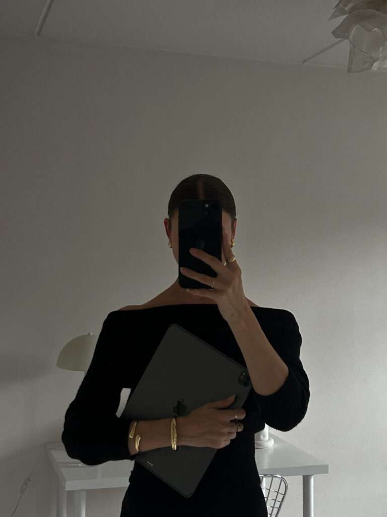 Image of a woman taking a photo of herself, wearing a black off the shoulder shirt, and holding an ipad.
