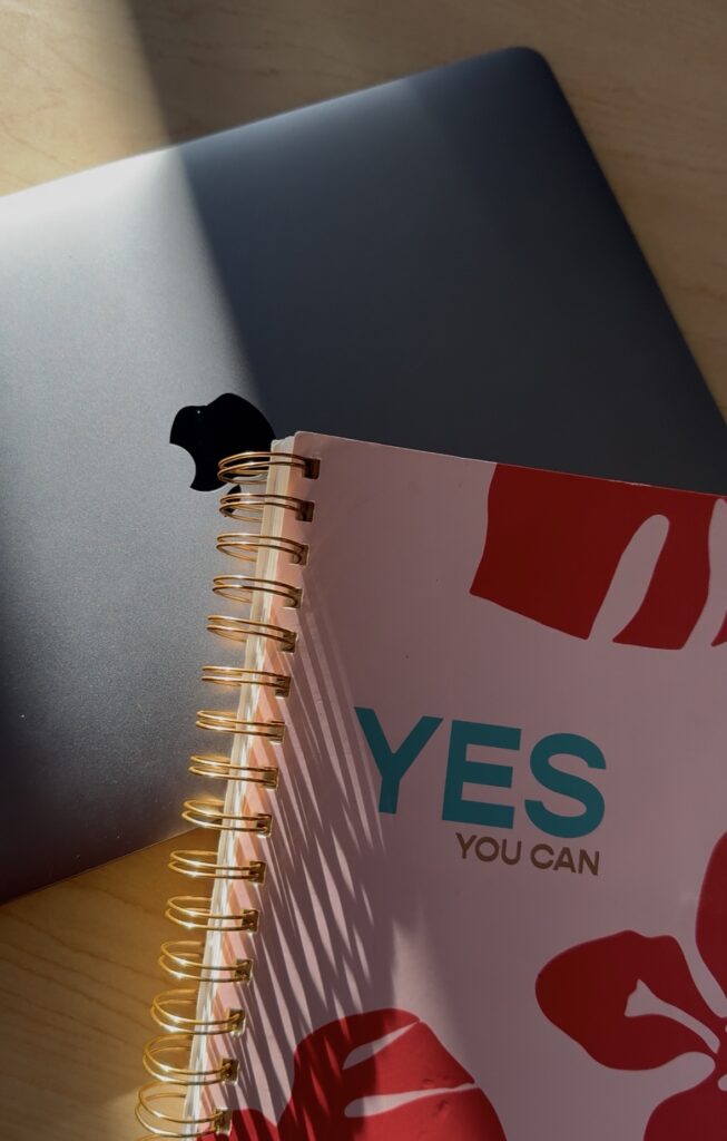 Image of a notebook that says, "Yes You Can" and a Macbook on a wooden table.