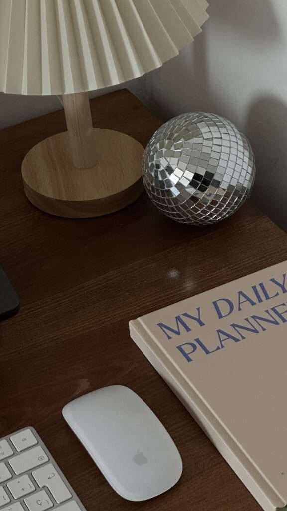 Image of a daily planner, apple mouse and keyboard, a desk lamp, disco ball on a brown wooden desk.